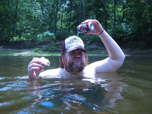 A burly bearded man wearing a trucker hat and no shirt is dousing himself with beer while partially submerged in a river. In his other hand is a partially eaten sandwich. Eww.