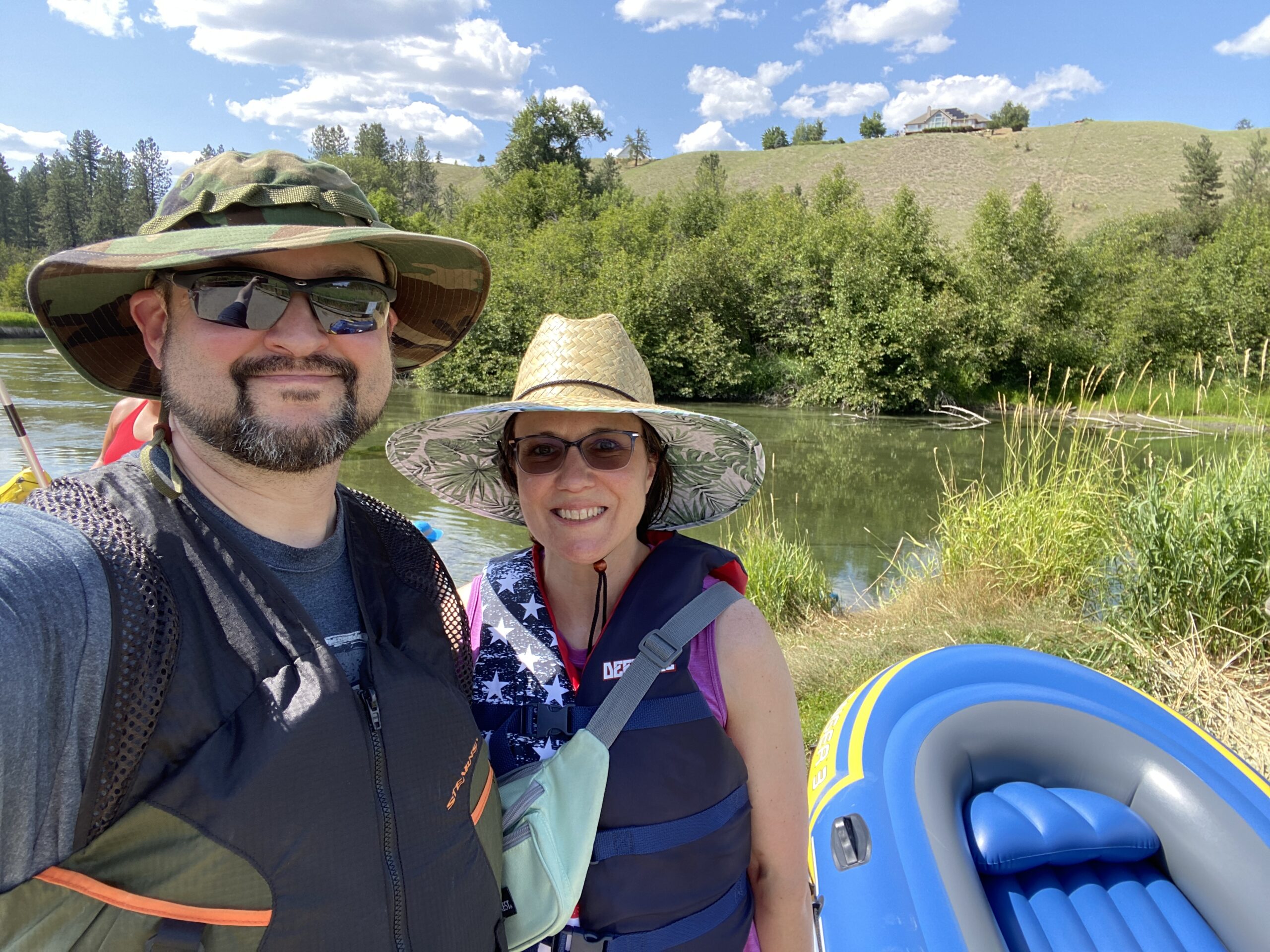 Erich Ebel and his wife smile at the camera with the Little Spokane River in the background and an inflatable blue raft at their feet.