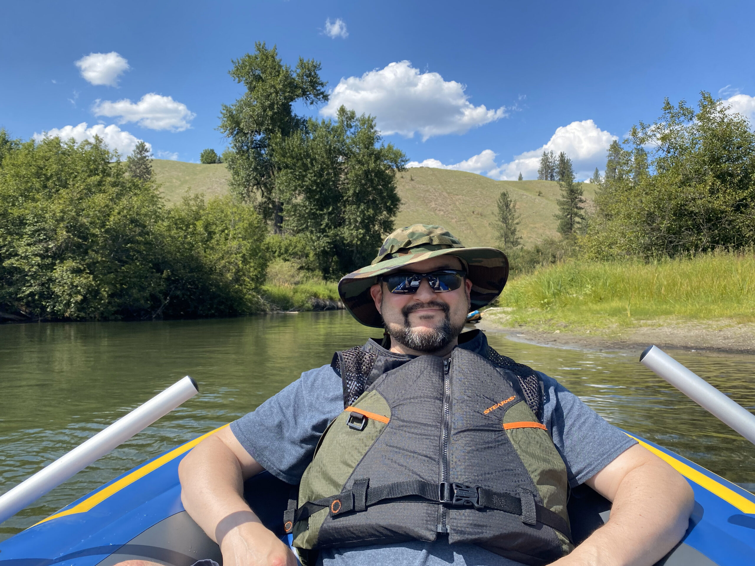 Erich Ebel smiling and reclining in an inflatable raft wearing a sun hat, sunglasses, and a life vest with the Little Spokane River and countryside in the background.